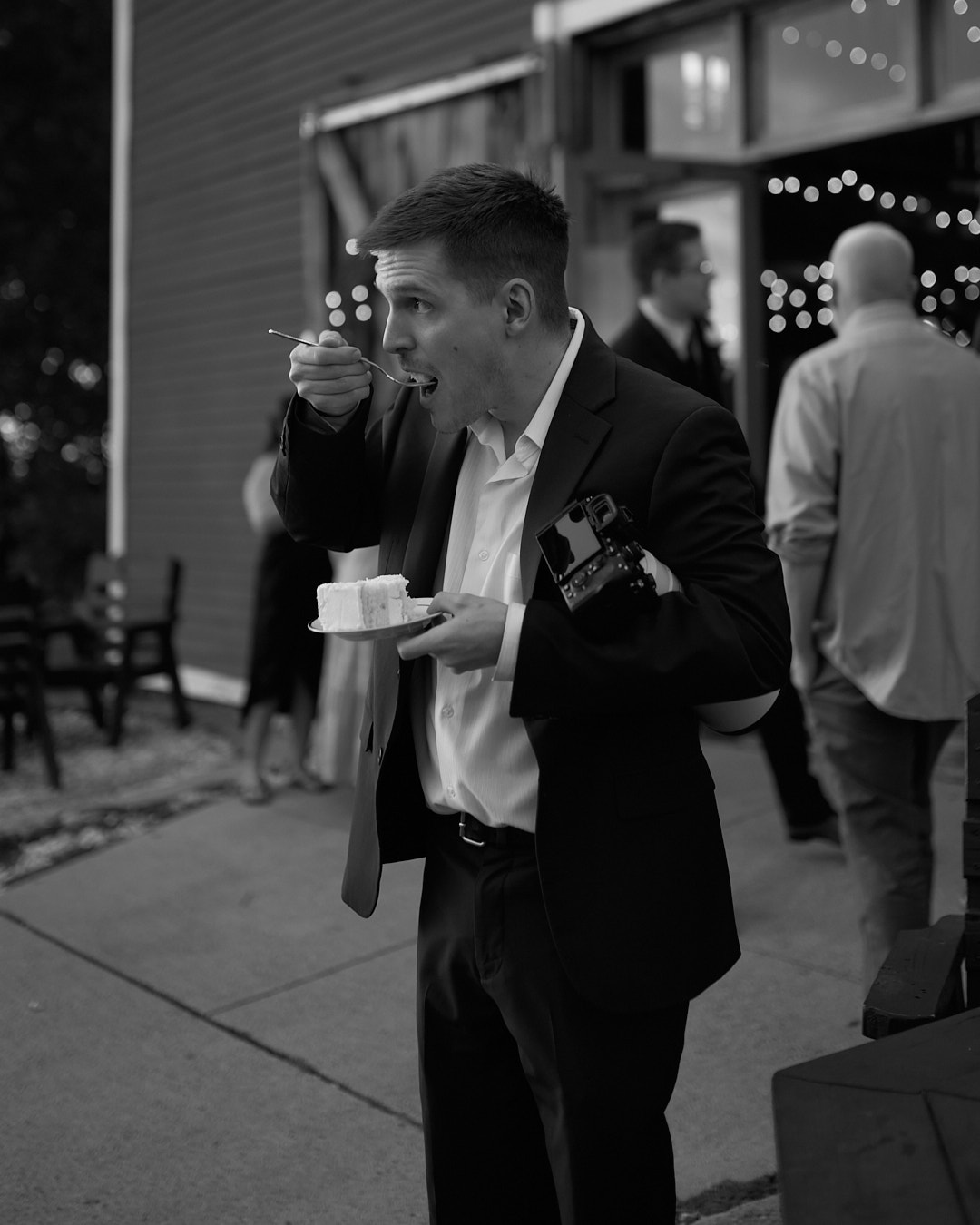 Cam of Two Friends Photography in a dark suit eating wedding cake, a camera tucked under his arm at an evening reception