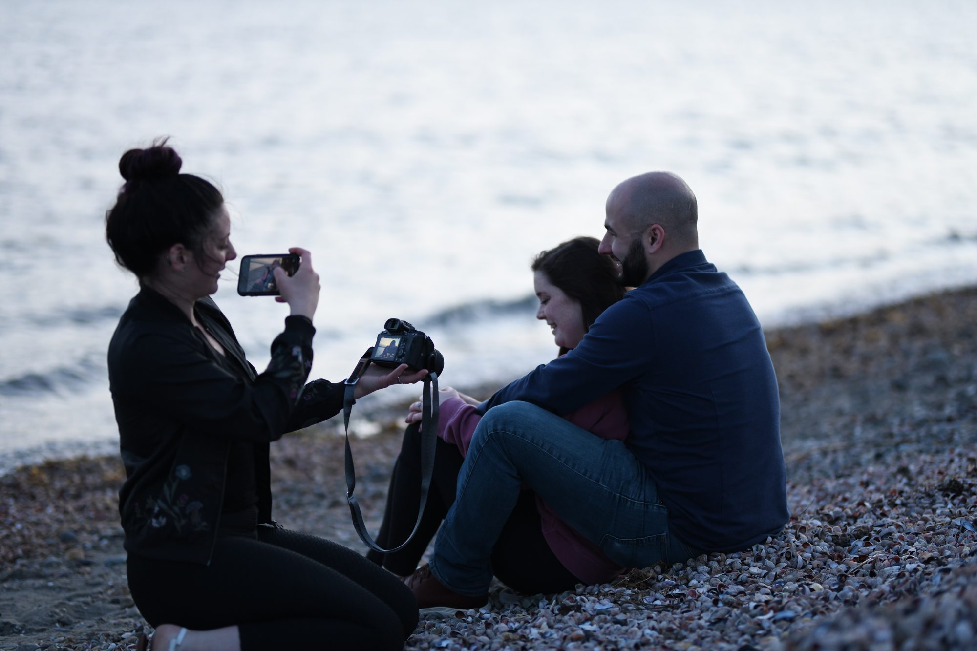 Maggie of Two Friends Photography photographing a couple sitting close at the water's edge on a New England beach at dusk